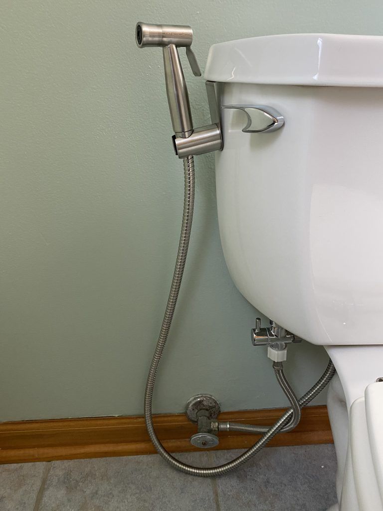 Hand held bidet installed and hanging in holder on toilet tank.