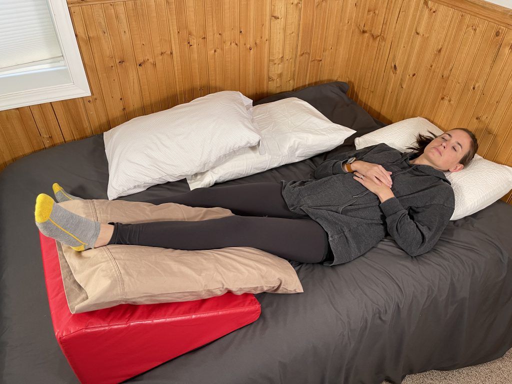 How to Sleep After Hip Replacement - woman is resting in bed on her back. Her left leg is resting on a pillow which is on a red wedge block.