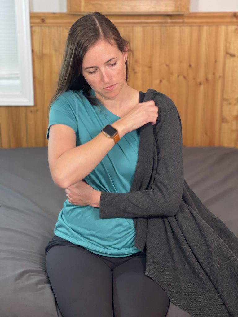 Woman sitting on the edge of a bed has her left arm across her abdomen. She is putting a sweater. Her right arm is pulling a sweater up to her left shoulder. The sweater is on her left arm that is across her abdomen.
