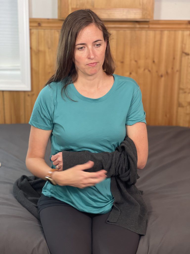 Woman sitting on the edge of a bed has her left arm across her abdomen. There is a sweater on the arm on her abdomen. The sweater is only on up to her elbow.