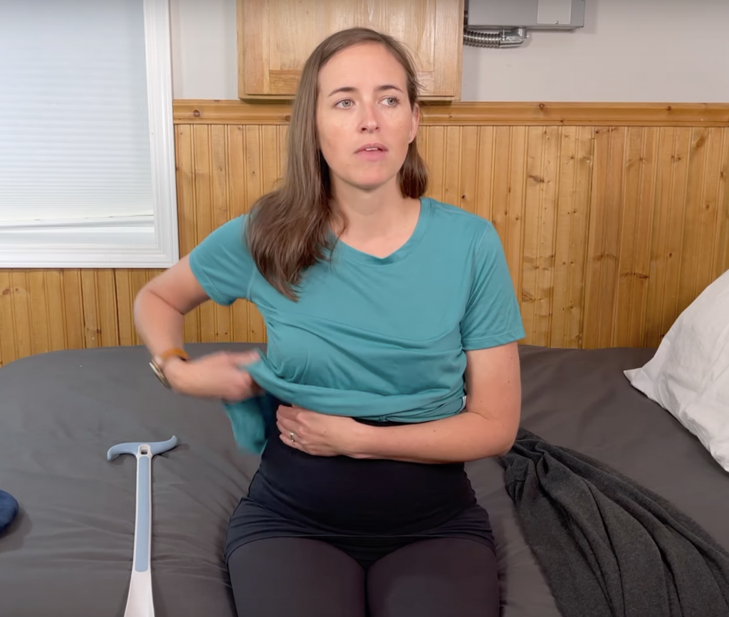 Woman sitting on the edge of a bed has her left arm across her abdomen. In her right hand, she has her shirt and she is bunching it up above her left arm which is across her abdomen.