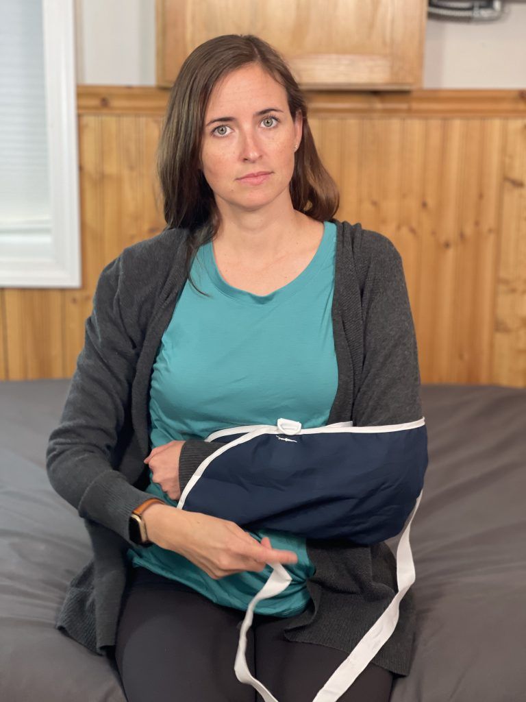 A woman is sitting up on the edge of a bed. Her left arm is in a sling and she is removing the shoulder strap with her right hand.