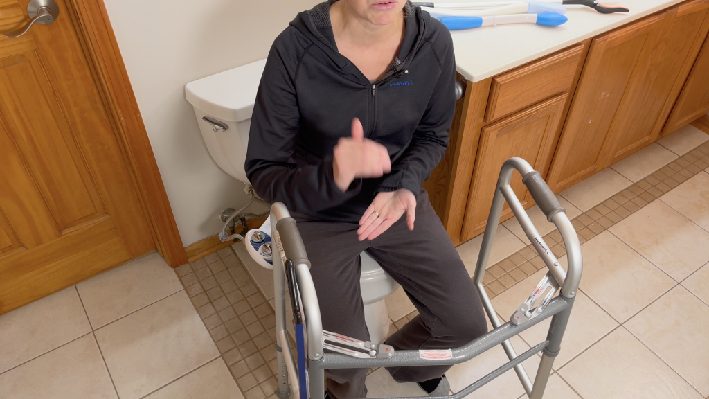 The image shows an adult woman sitting on a low toilet with the lid closed, with a walker in front of her. She is wearing a black zip-up jacket and black pants, and appears to be in the middle of a conversation or explaining something, as she is gesturing with her right thumb upwards.