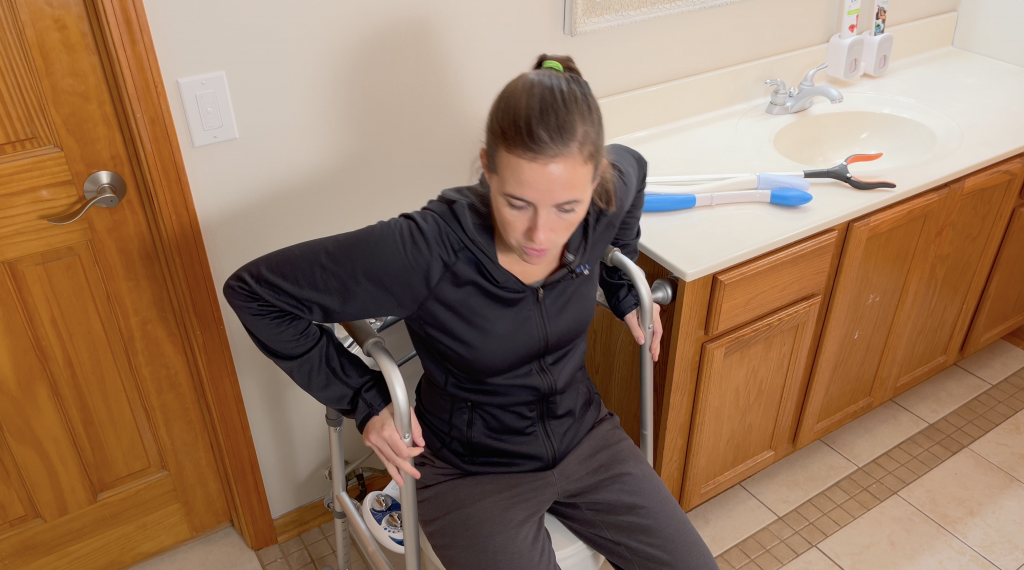 A woman is in the process of standing up from a closed toilet seat using a walker for assistance. She is wearing a black zip-up jacket and dark pants, with her hair pulled back into a ponytail. Her focus is downward, possibly concentrating on her movement. Her arms are pushing down on the bracing of the walker. To the right is a bathroom vanity with a white sink and faucet, where two long-handled reachers are laid out. The room has tan floor tiles and a wooden door, indicating a residential bathroom designed with accessibility in mind.