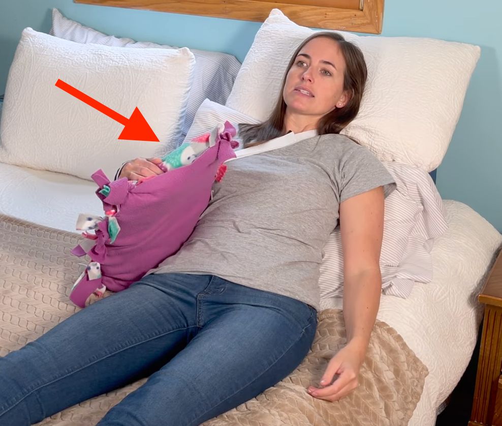 How to Sleep After Shoulder Surgery - woman in sling laying in bed on her back with red arrow pointing to her injured arm that is holding a pillow off to the side of her body