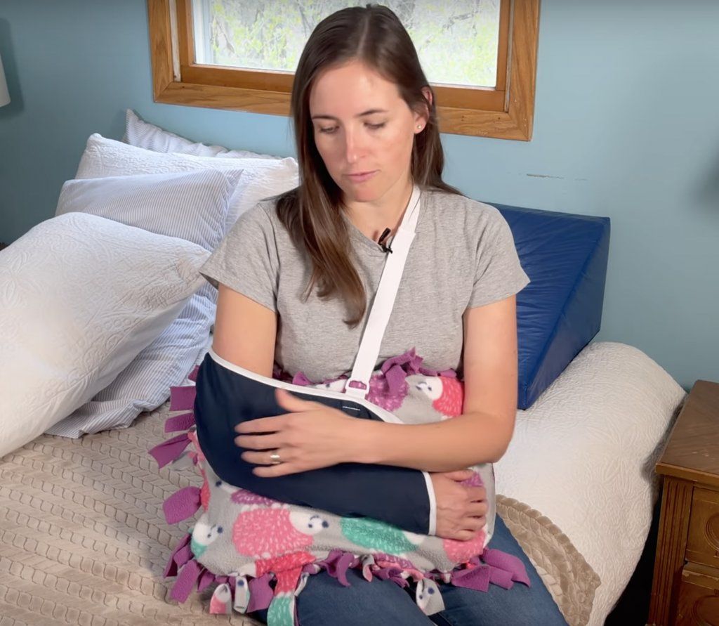 How to Sleep After Shoulder Surgery - a woman with her right arm in a sling sitting on a bed. A small pillow is between her arm in the sling and her body.