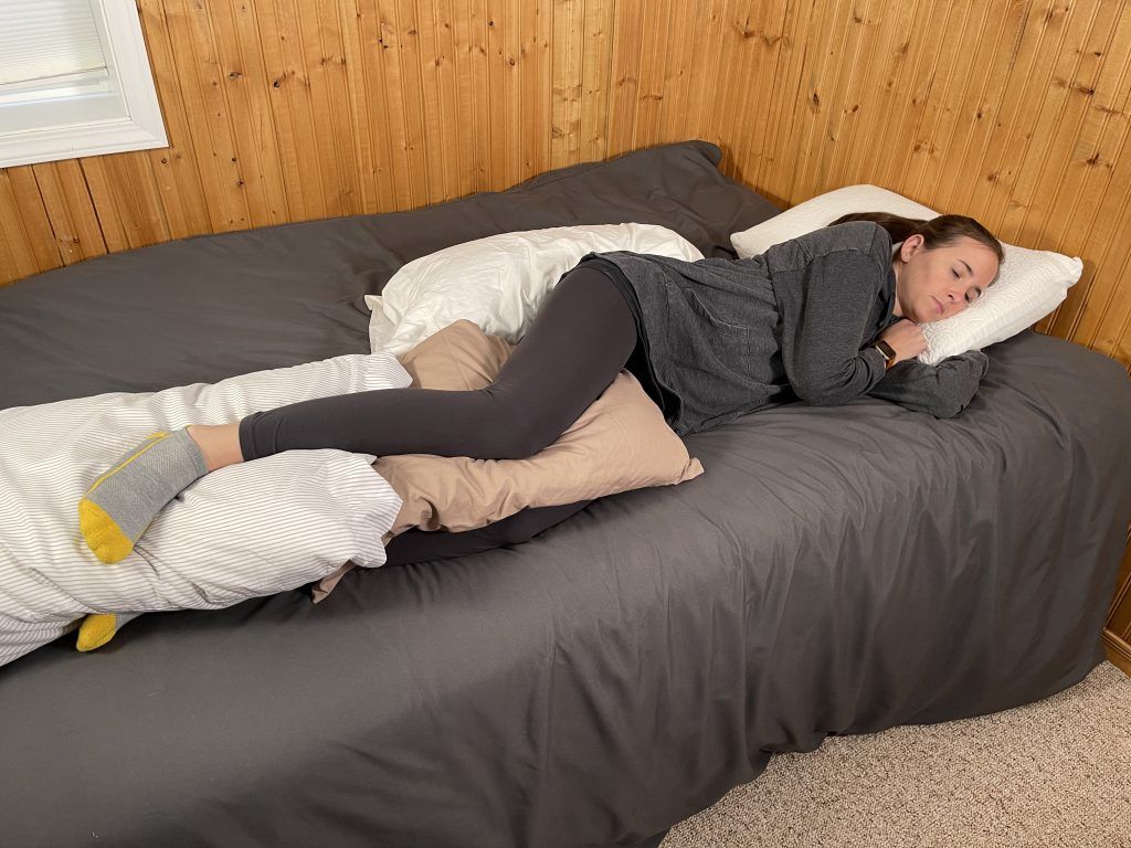 How to Sleep After Hip Replacement - woman is sleeping in bed on her side. She is sleeping on her left side with her head on a pillow, two pillows in between her legs, and a pillow behind her.
