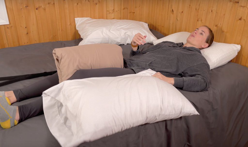 How to Sleep After Hip Replacement - woman laying down on her back in a bed with her legs extended out. There is a pillow between her knees and she has another pillow in her left hand next to her left leg.