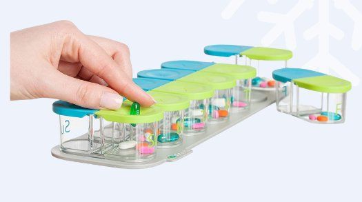 A green and blue pill organizer set on top of a magnetic gray base. A hand is inserting pills through the top of the organizer lid.