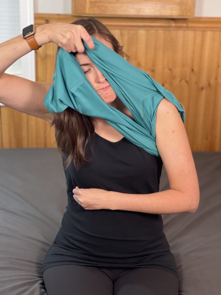 Woman sitting on the edge of a bed has her left arm across her abdomen. She is putting a t-shirt. Her left arm is through and the shirt is on her left shoulder and with her right hand, she is pulling the t-shirt over her head.