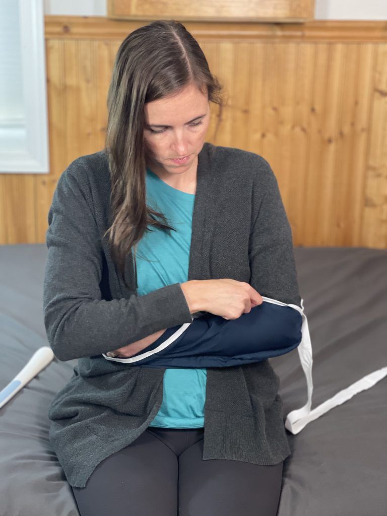 Woman sitting on the edge of a bed has her left arm across her abdomen. She has a sweater on and with her right hand is putting a sling on her left arm.