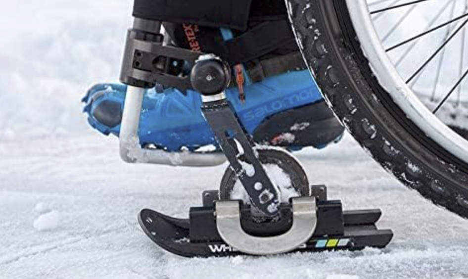 Close-up image featuring a wheelchair wheel with a specialized ice grip attachment that looks like a miniature snow ski on snowy ground. The grip device is clamped onto the wheelchair's front caster. Snow and ice particles are visible on the wheel and the grip, indicating recent use. The device is part of an adaptive technology to improve the wheelchair's maneuverability in winter conditions. How to Avoid Slips and Falls on Ice: Essential Winter Safety Tips