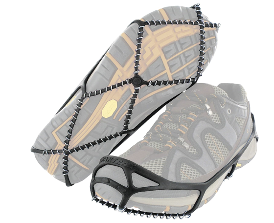 Image displaying a pair of grey hiking shoes with attached black traction cleats. The cleats are fitted snugly over the soles of the shoes, connected by a series of elastic bands ensuring a tight grip. The soles of the shoes have a yellow and grey tread pattern, and the cleats feature multiple small, black spikes arranged in rows designed to increase grip on slippery surfaces. The cleats are likely used to provide additional traction on ice or snow.