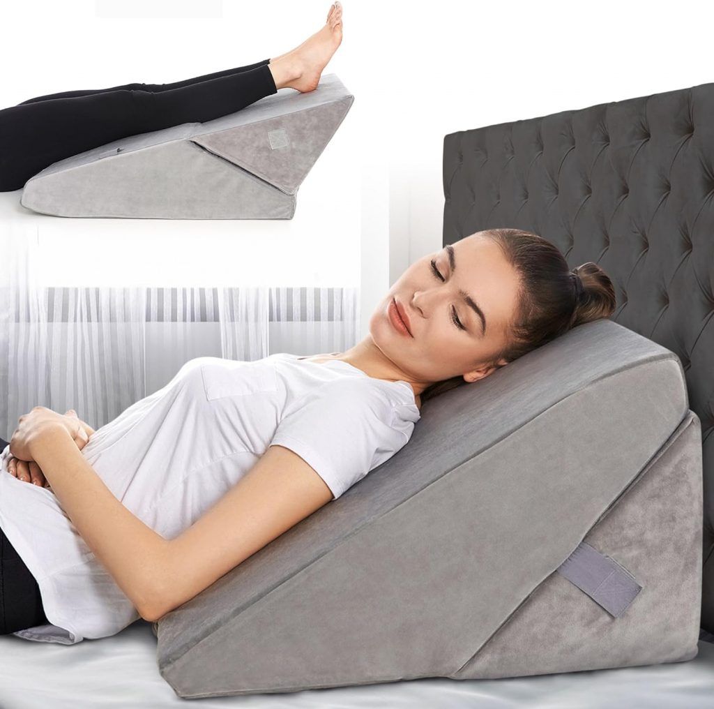 A woman resting comfortably on a large gray wedge pillow designed to elevate the upper body. She is lying on her back with her hands folded on her abdomen and her eyes closed, indicating relaxation. Her head is at the higher end of the pillow, which supports her back and neck. In the inset image at the top left corner, the same wedge pillow is shown being used to elevate the woman's legs, which are extended upward. How to Sleep with Rib Injury.