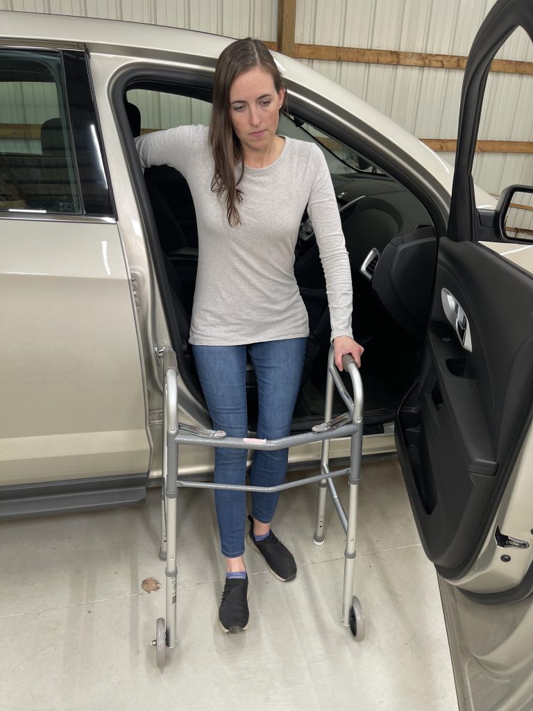 A woman uses a walker for support while reaching back to the car. She holds the car door frame with one hand and the walker with the other, demonstrating a safe technique for entering a vehicle with mobility assistance. She is wearing a light gray long-sleeve shirt, blue jeans, and black slip-on shoes, with the car door open and the interior partially visible. How to Get In and Out of the Car After Hip Replacement